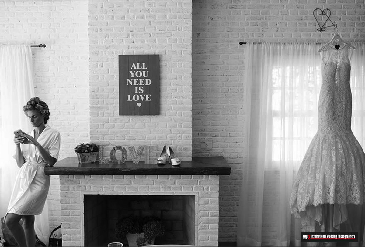 Noiva se conecta ao celular, ao lado o vestido dela esta pendurado na janela a espera dela para o casamento.